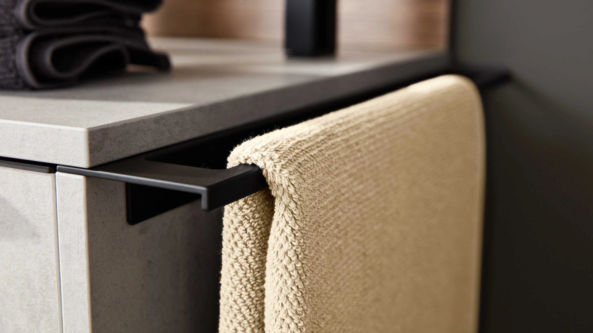 Beiges Handtuch in Nahaufnahme auf schwarzer Halterung vor grauem Badmöbel.