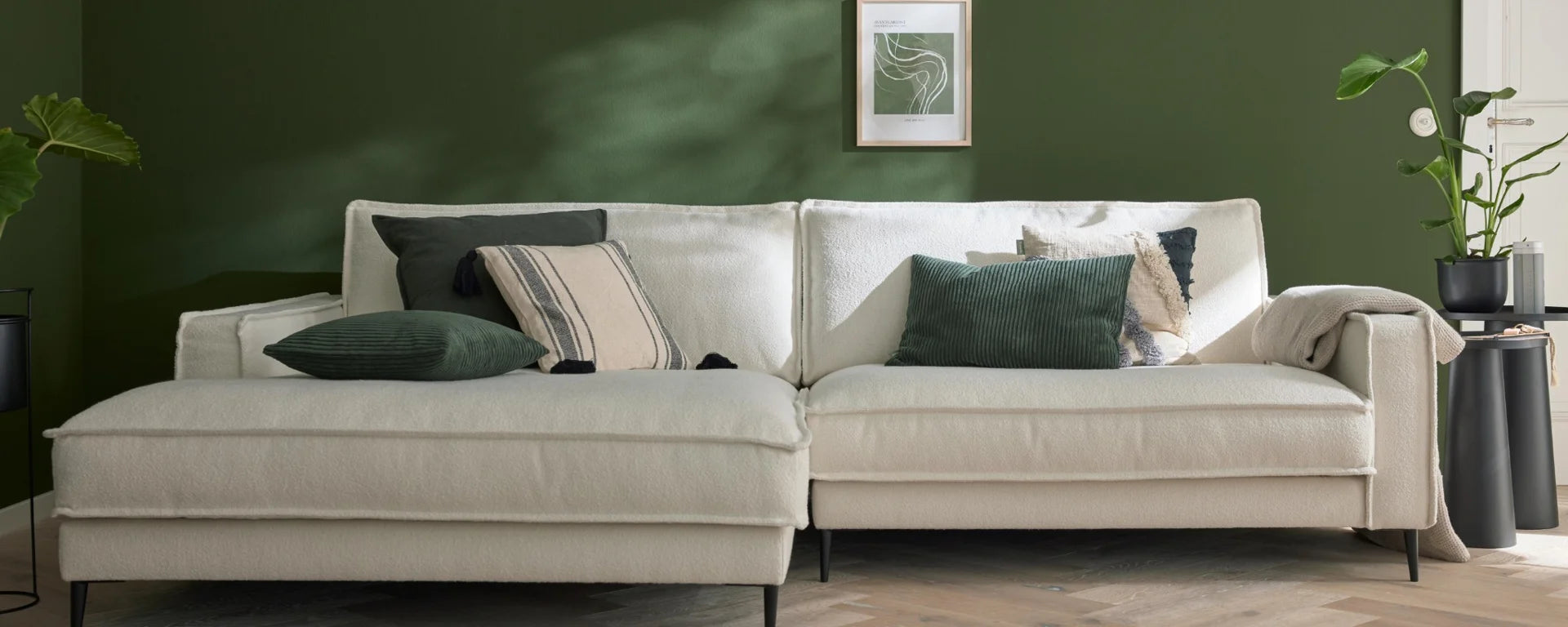 Beiges Sofa mit grünen Kissen vor dunkelgrüner Wand in modernem Wohnzimmer mit Pflanzen.